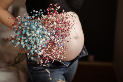 Gravid kvinna i jeans och bh håller en bukett med färgad brudslöja