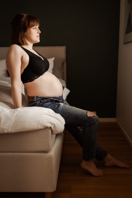 Gravid kvinna sitter i profil på en säng med bh och jeans