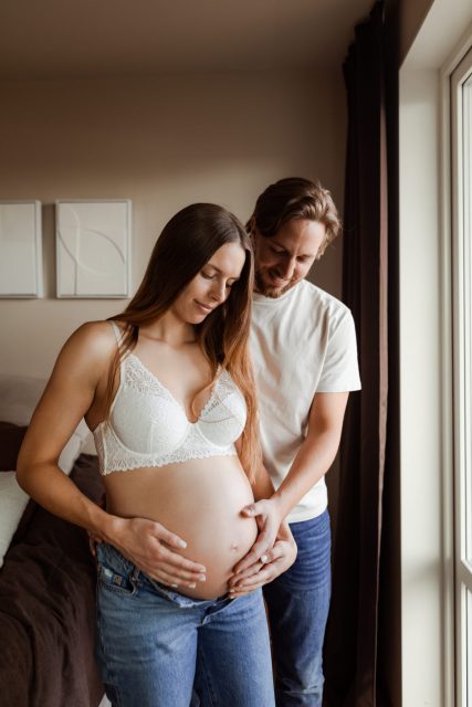 En gravid kvinna står med jeans och bh framför ett fönster medan hennes man håller en hand på hennes gravidmage