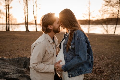 En gravid kvinna fotograferas i solnedgången tillsammans med sin partner, gnuggar näsorna och skrattar och håller en hand på magen.