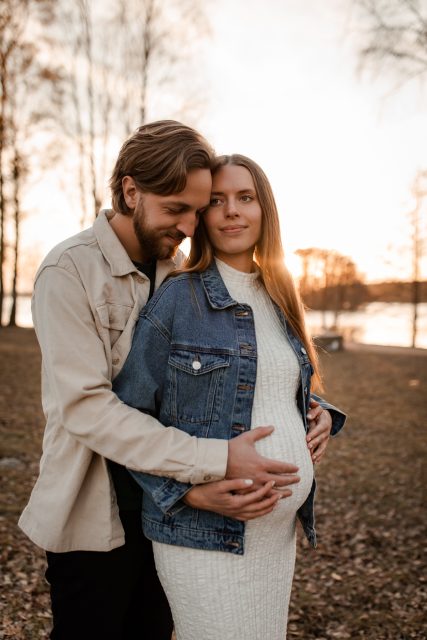 En gravid kvinna fotograferas i solnedgången tillsammans med sin partner som håller om kvinnan och har sin hand på hennes mage.