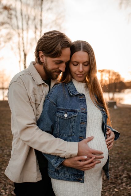 En gravid kvinna fotograferas i solnedgången tillsammans med sin partner som håller om kvinnan och har sin hand på hennes mage.