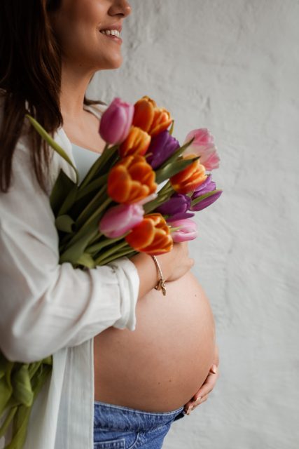 Gravid kvinna i profil, klädd i jeans och vit skjorta, står med ena handen på magen och håller i en bukett tulpaner i andra handen.