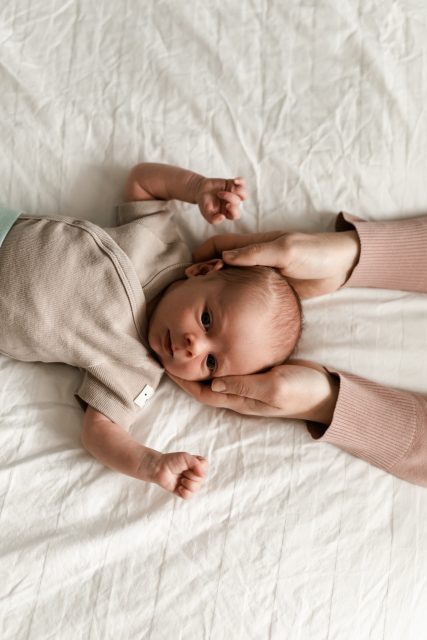 Nyfödd bebis ligger vaken på en säng med sin mammas händer om huvudet.