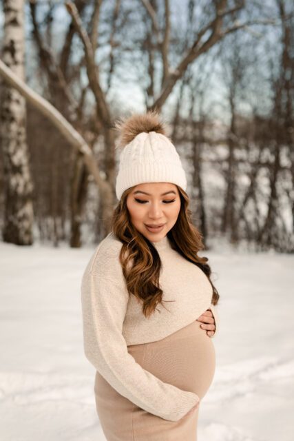 Gravid kvinna står ute i snön iklädd beiga kläder och mössa på sin gravidfotografering i Småland