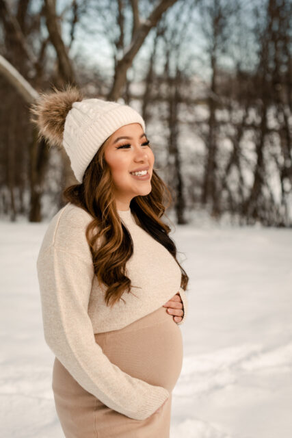 Gravid kvinna står ute i snön iklädd beiga kläder och mössa på sin gravidfotografering i Småland